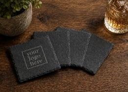 Stack of square black slate coasters with an engraved logo panel on a wood table